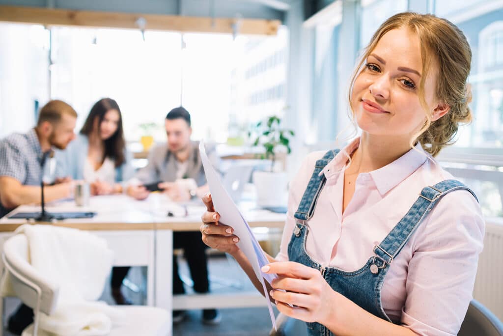 Formation en marketing : jeune étudiante en alternance au premier plan, tenant des documents, ses collègues sont assis autour d'un bureau en arrière-plan et travaillent ensemble